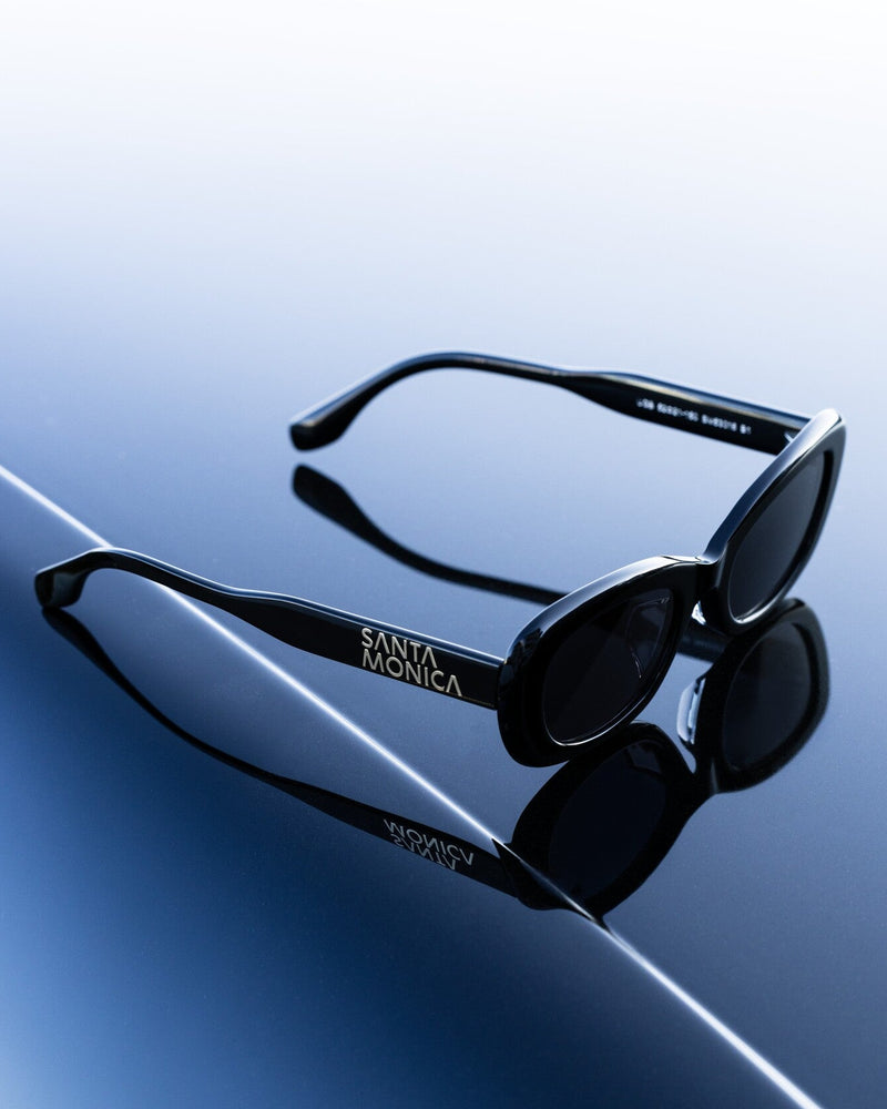 Black sunglasses with 'Santa Monica' branding on a reflective blue surface